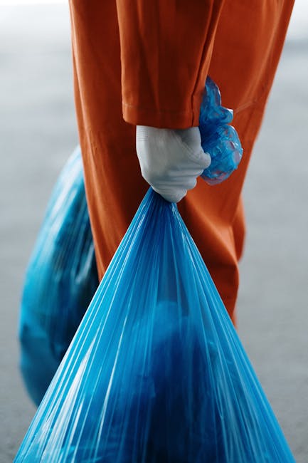 Close-up of a person wearing an orange protective suit and a white glove, holding a blue plastic bag filled with waste or packing materials, with the background blurred to focus on the individual's ha